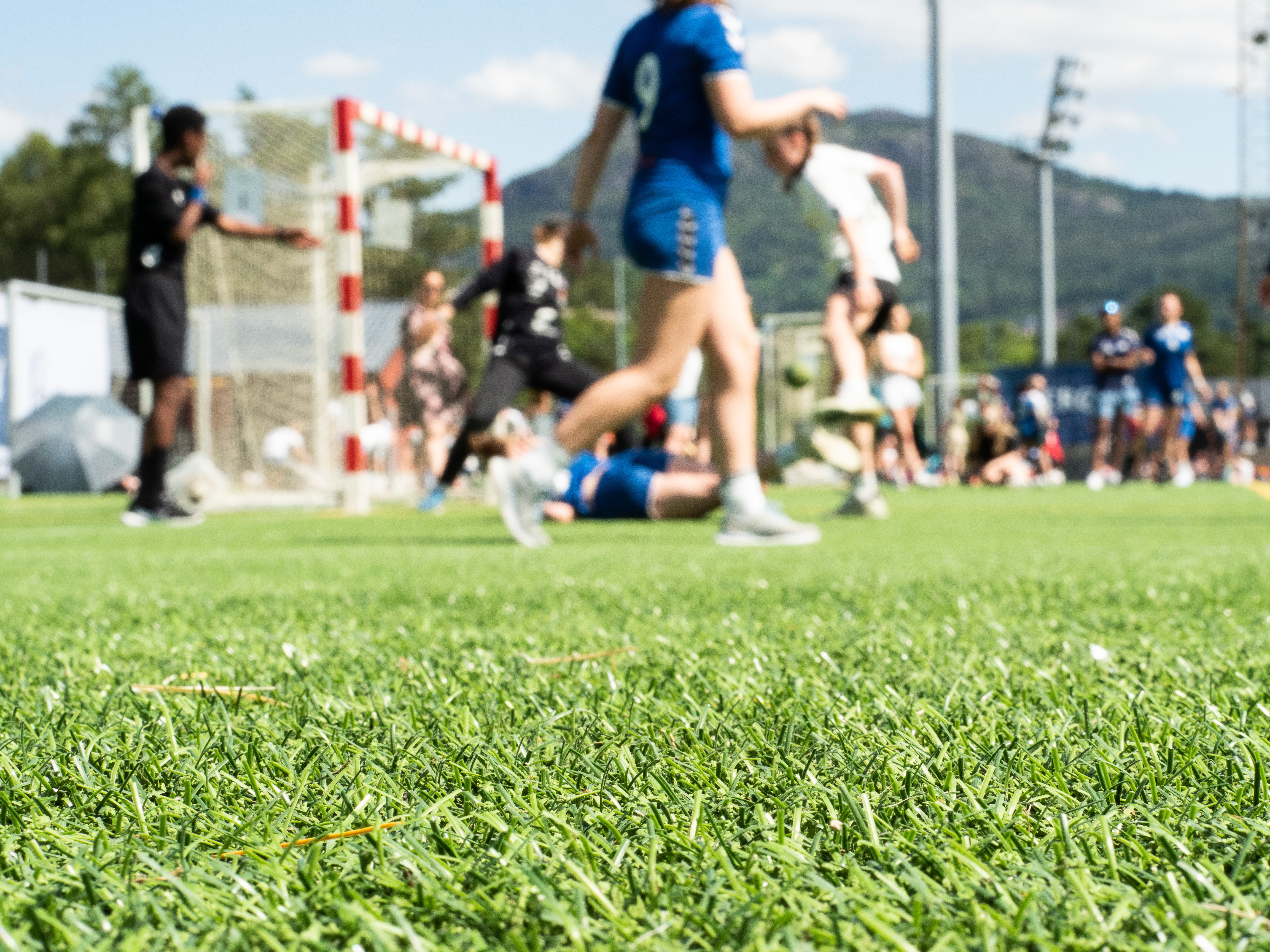 Grønt gress i fokus, flere håndballspillere spiller håndball i ufokus