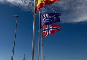 Fabrikkbygning sett forfra på skrå. Den er plassert midt i bildet på sandgrus med himmel og skyer bak.