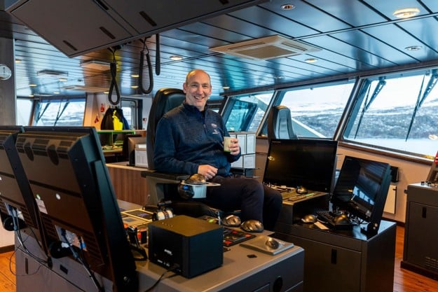 En smilende mann sitter i en stol i styrerommet på en tråler, rundt han er vinduer med utsikt til havet rundt ham.