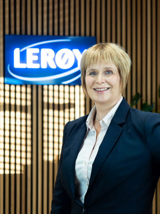 En smilende glad kvinne med hvit skjorte og blå blazer, bak henne lyser et skilt med teksten "Lerøy" opp på spileveggen.