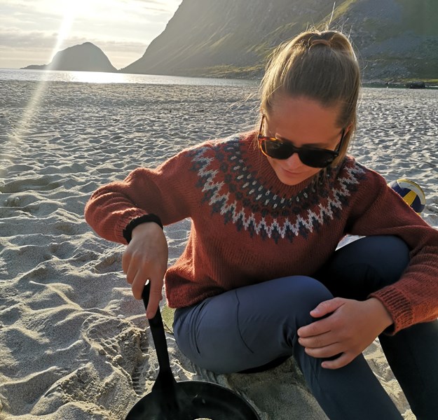 Astrid on a beach with a mountain behind her. She is wearing a wool sweater and is frying a pancake in a frying pan.