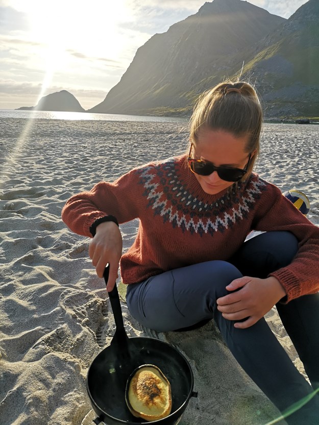 Astrid på en strand med fjell i bakgrunnen. Hun har på seg en ullgenser og steker en pannekake i en stekepanne.