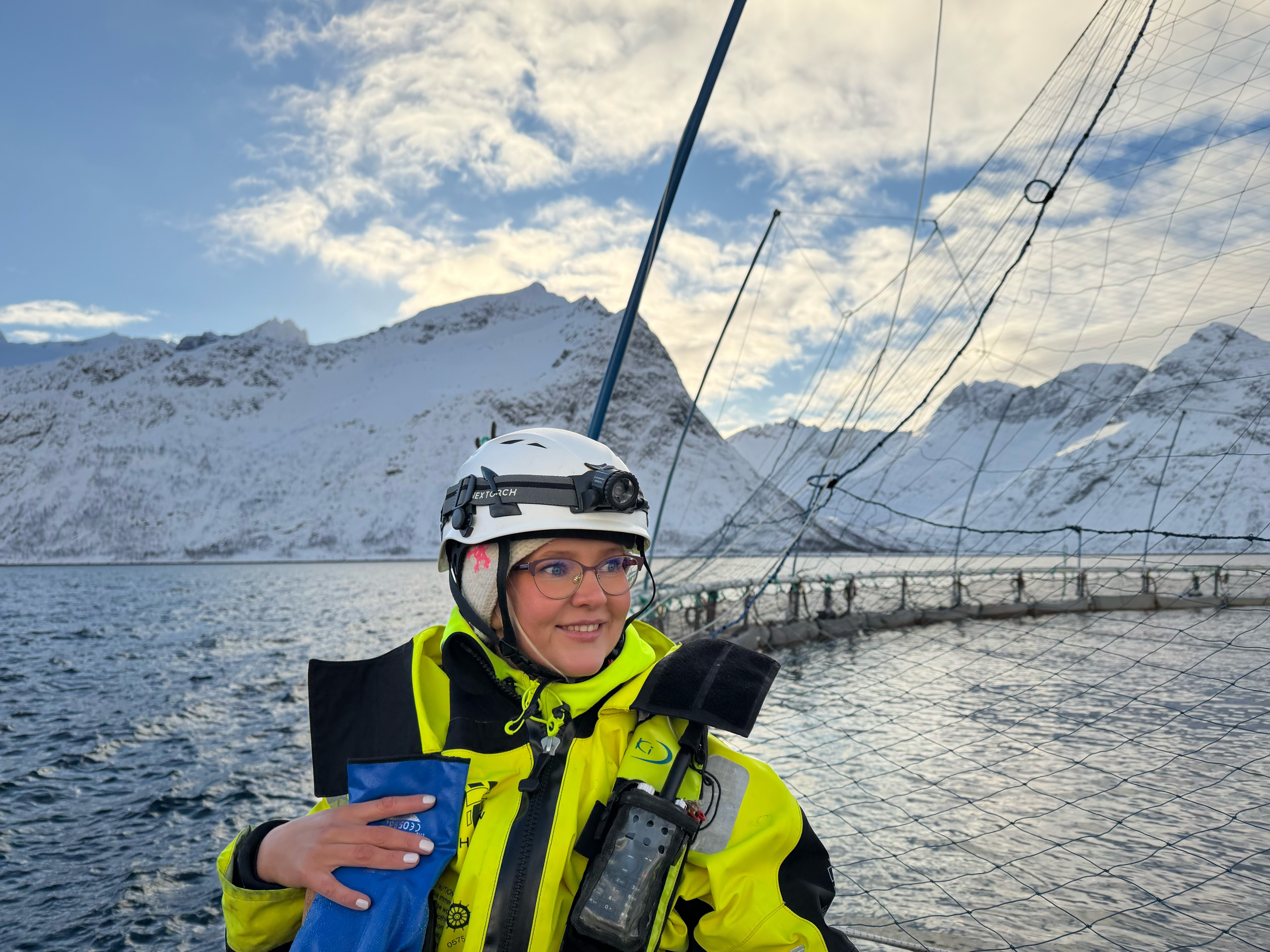 Ung kvinne ser mot høyre, hun er ikledd hjelm og arbeidsklær, bak henne ser man en merd og fjell.