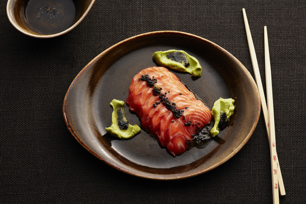 Mørkt fat med oransje-røde sashimi biter som lyser opp fatet. Rundt er grønne klatter sterkt 