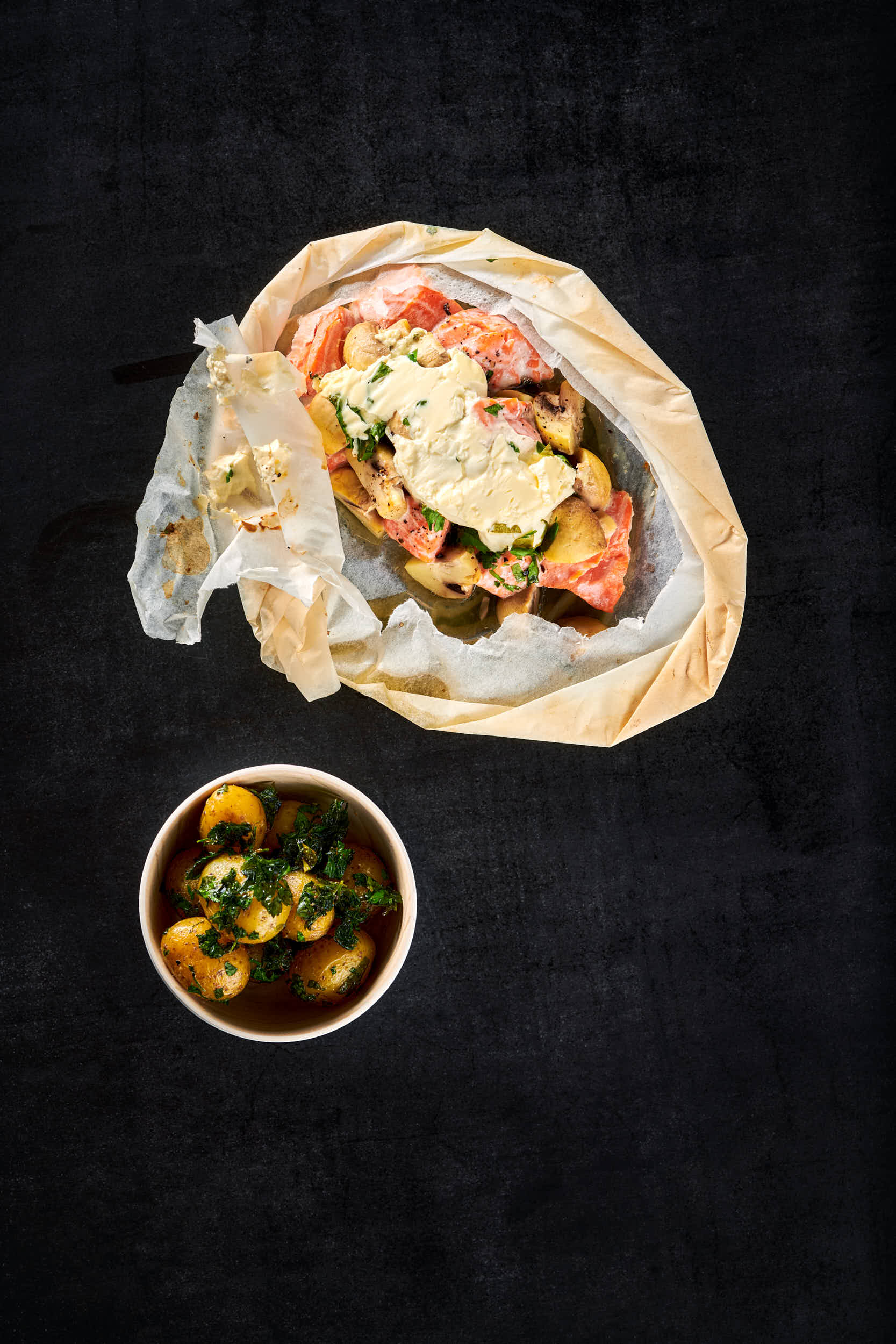 Mørk bakgrunn, en pakke av bakepapir med ørret og grønnsaker, en skål med saus like ved. 