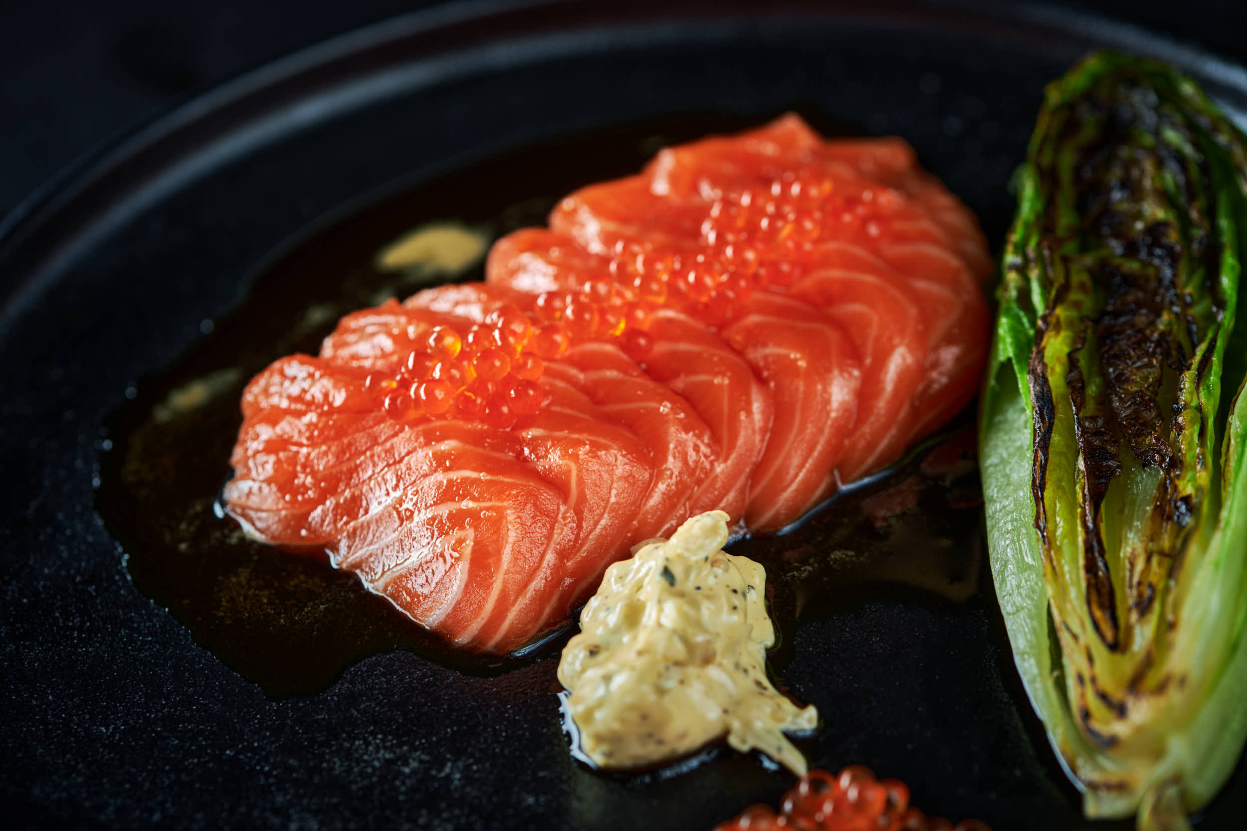 Sashimi av ørretloin med grillet hjertesalat og peppermajones dandert fint på et svart fat.