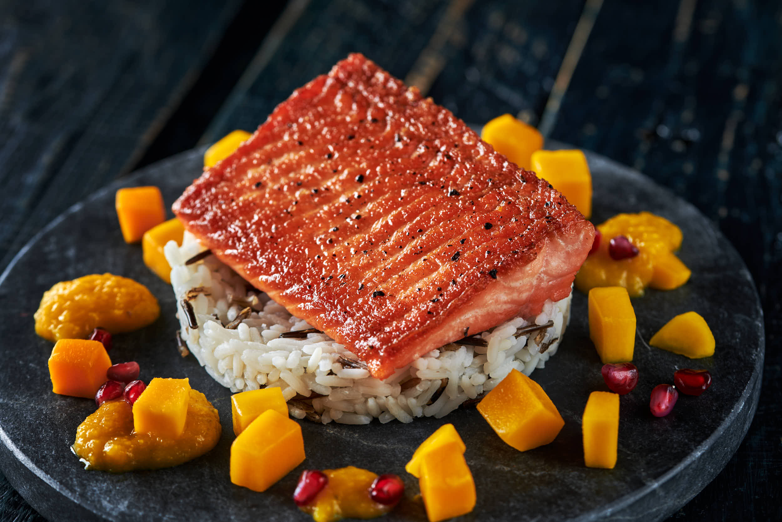 Svart bakgrunn og fat med villris med stekt ørretloin oppå og mangobiter rundt. 